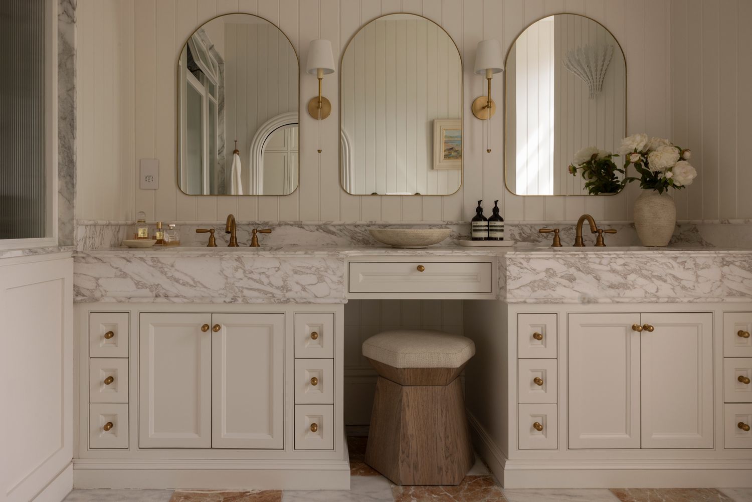 Elegant white marble bathroom with gold fixtures features a double vanity and large arched mirror in a luxurious design.