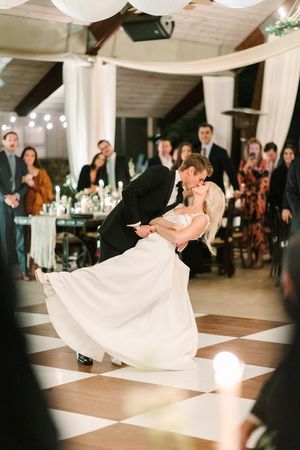 A couple shares a romantic dip during their first dance on a black and white checkered dance floor at an indoor reception.