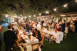 Candlelit outdoor wedding reception with string lights and long tables set up on a garden lawn at dusk.