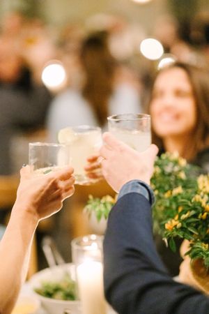 People raise glasses in a toast at a celebration dinner with soft bokeh lights and yellow flowers in the foreground.