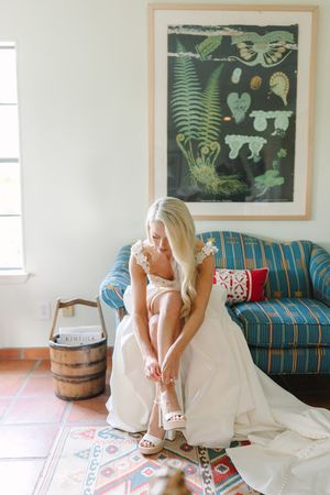 A bride in a white wedding dress sits on a patterned rug in front of a teal couch with botanical artwork on the wall.