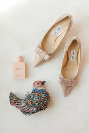 Blush pink bow-detailed Jimmy Choo heels with perfume and a decorative bird brooch photographed on a flatlay at San Antonio Wedding.