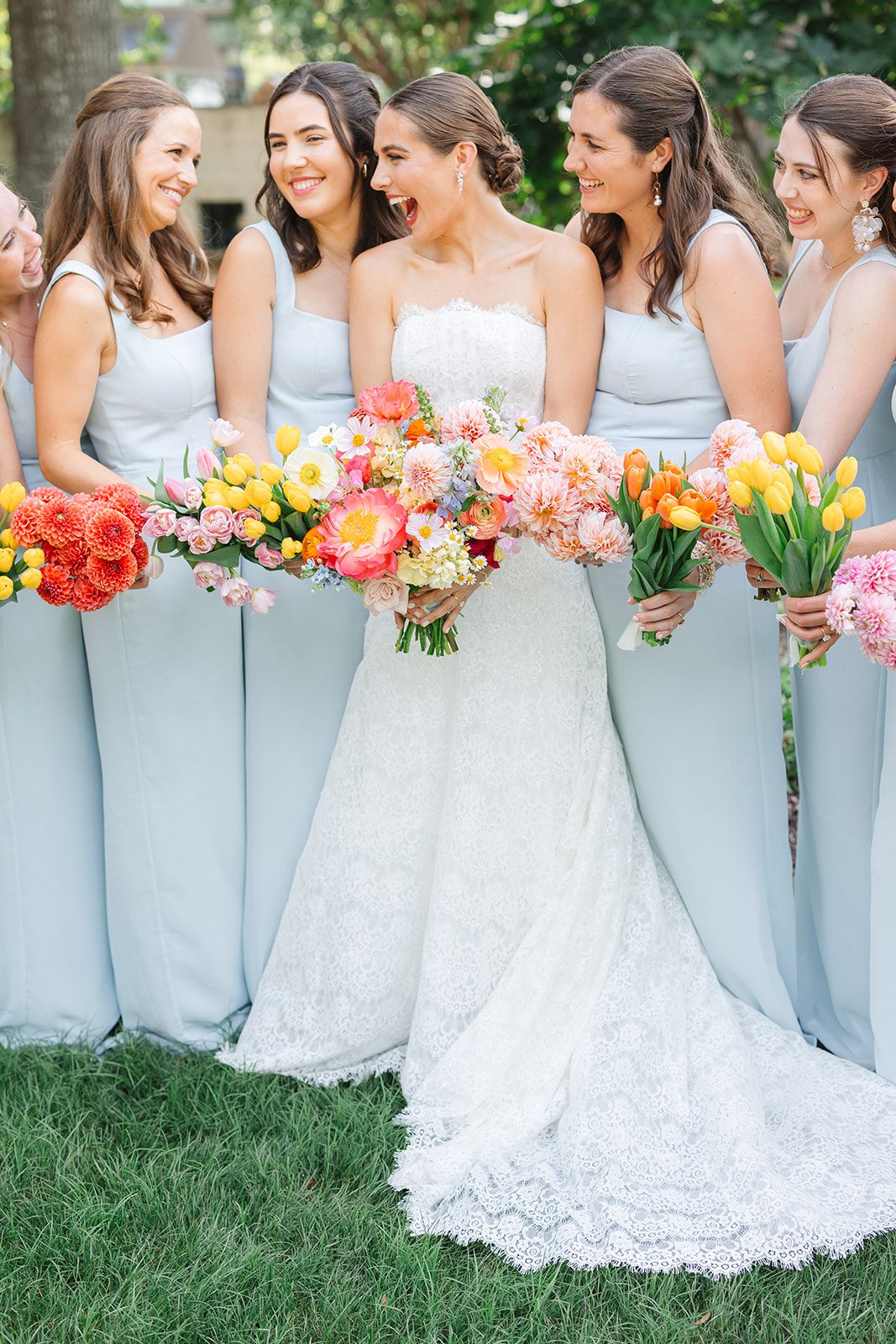 Bridesmaids in pale blue dresses holding vibrant mixed flower bouquets alongside bride in lace gown at the Dallas Arboretum's a Tasteful Pla