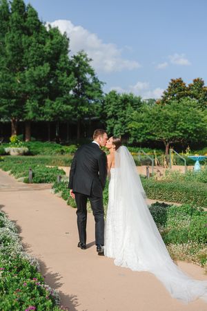 A couple in wedding attire walking on a garden path surrounded by lush greenery and flowers at the Dallas Arboretum's a Tasteful Place