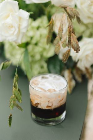 A layered coffee cocktail with cream foam and dried floral decorations in a rustic setting.