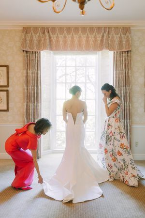 Three people help adjust a flowing white wedding gown by a bright window with elegant curtains.