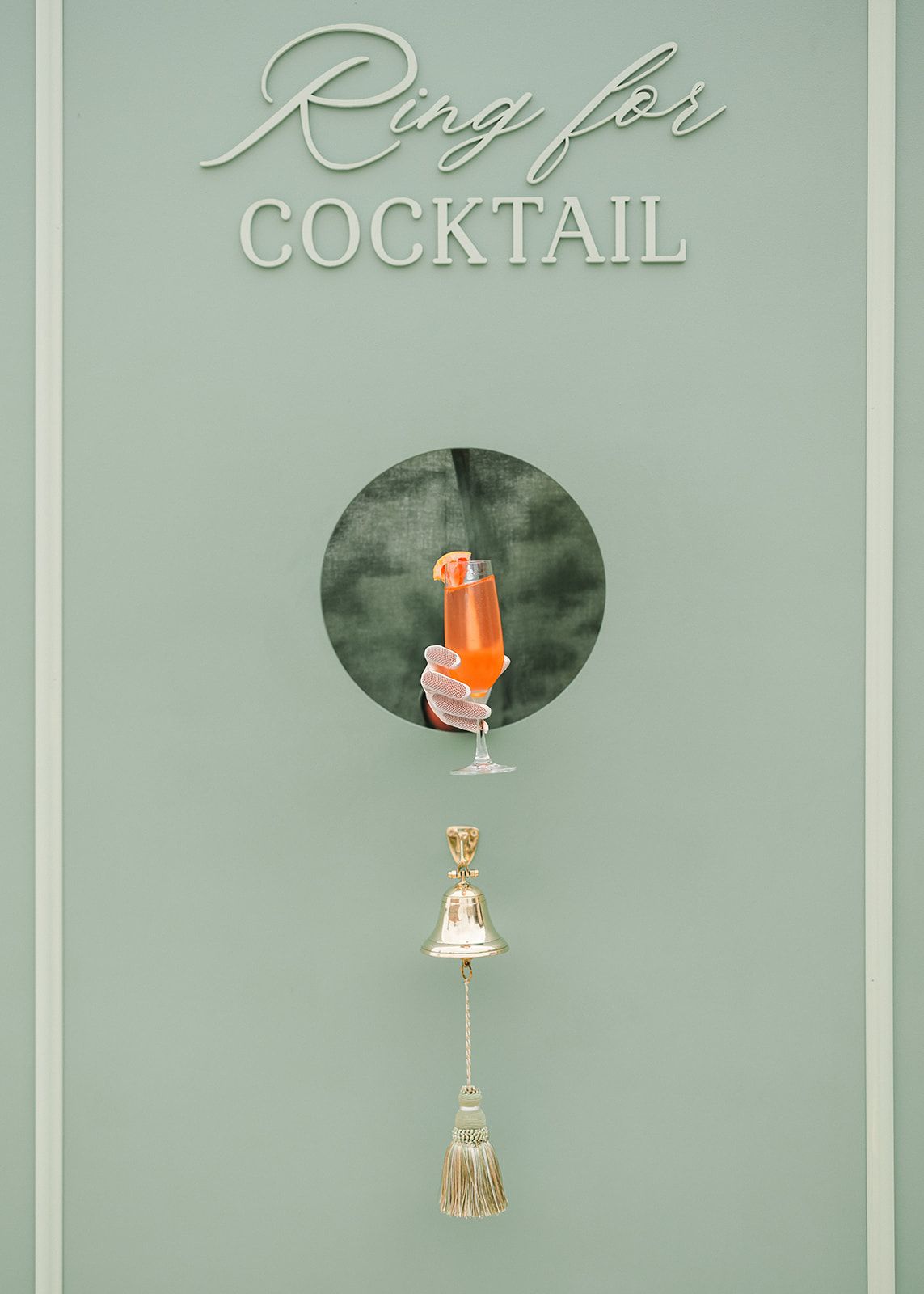 A minimalist sage green wall sign reading 'Ring for Cocktail' with a circular window showing an orange drink.