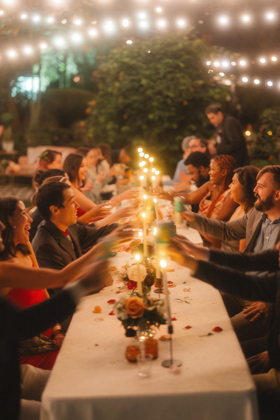 Guests gather around a long candlelit table at an evening outdoor dinner party with string lights overhead.