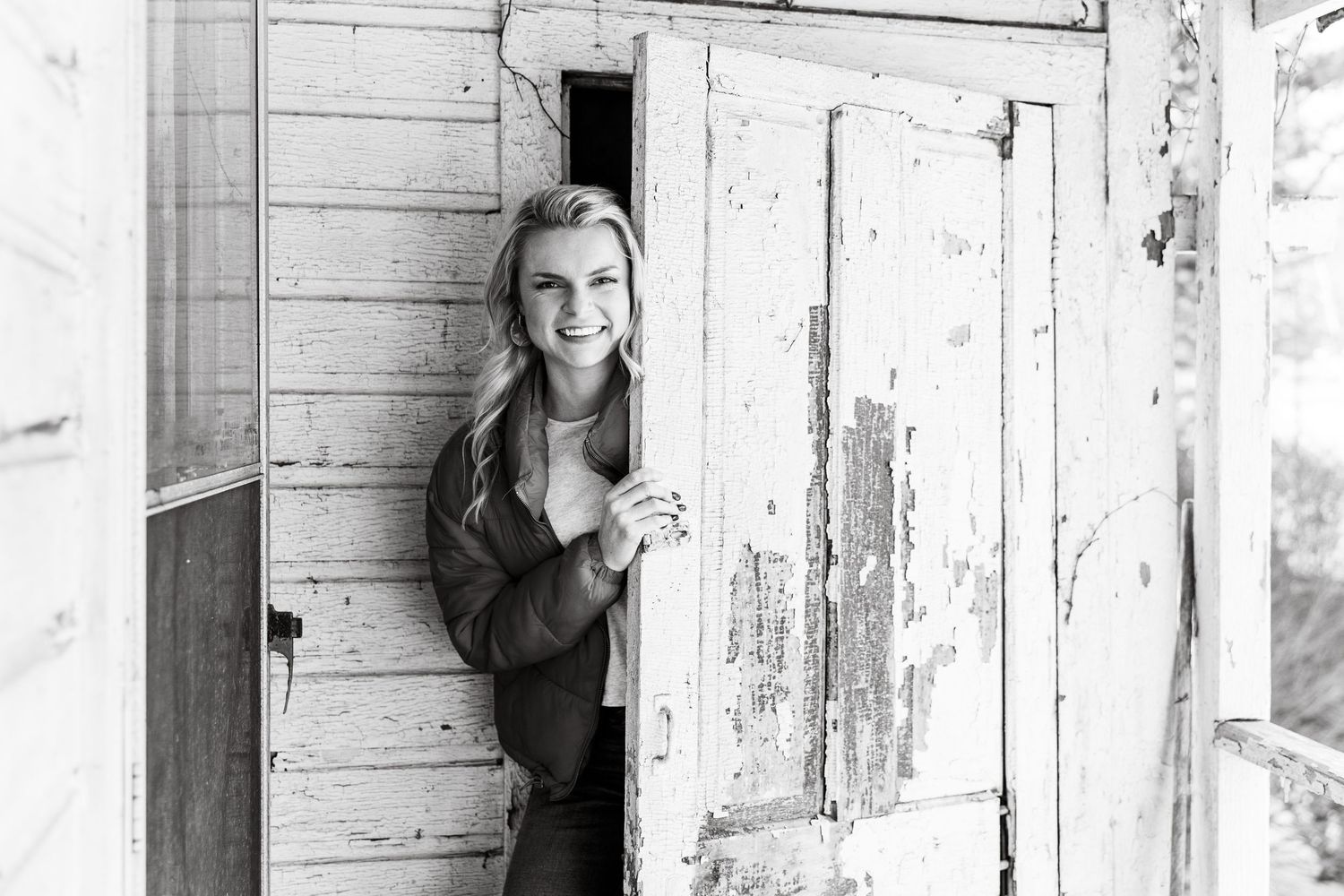 Black and white portrait of person peeking through rustic wooden doorway.