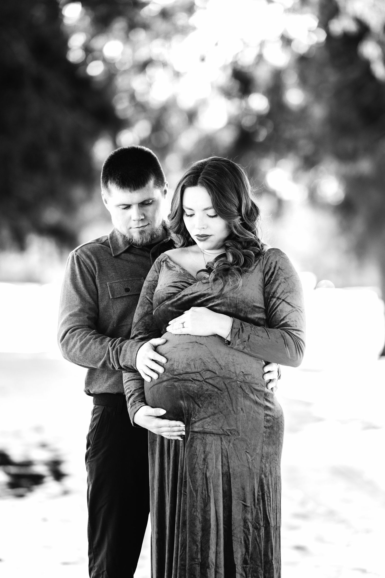 Black and white maternity photo of couple embracing outdoors with bokeh background.