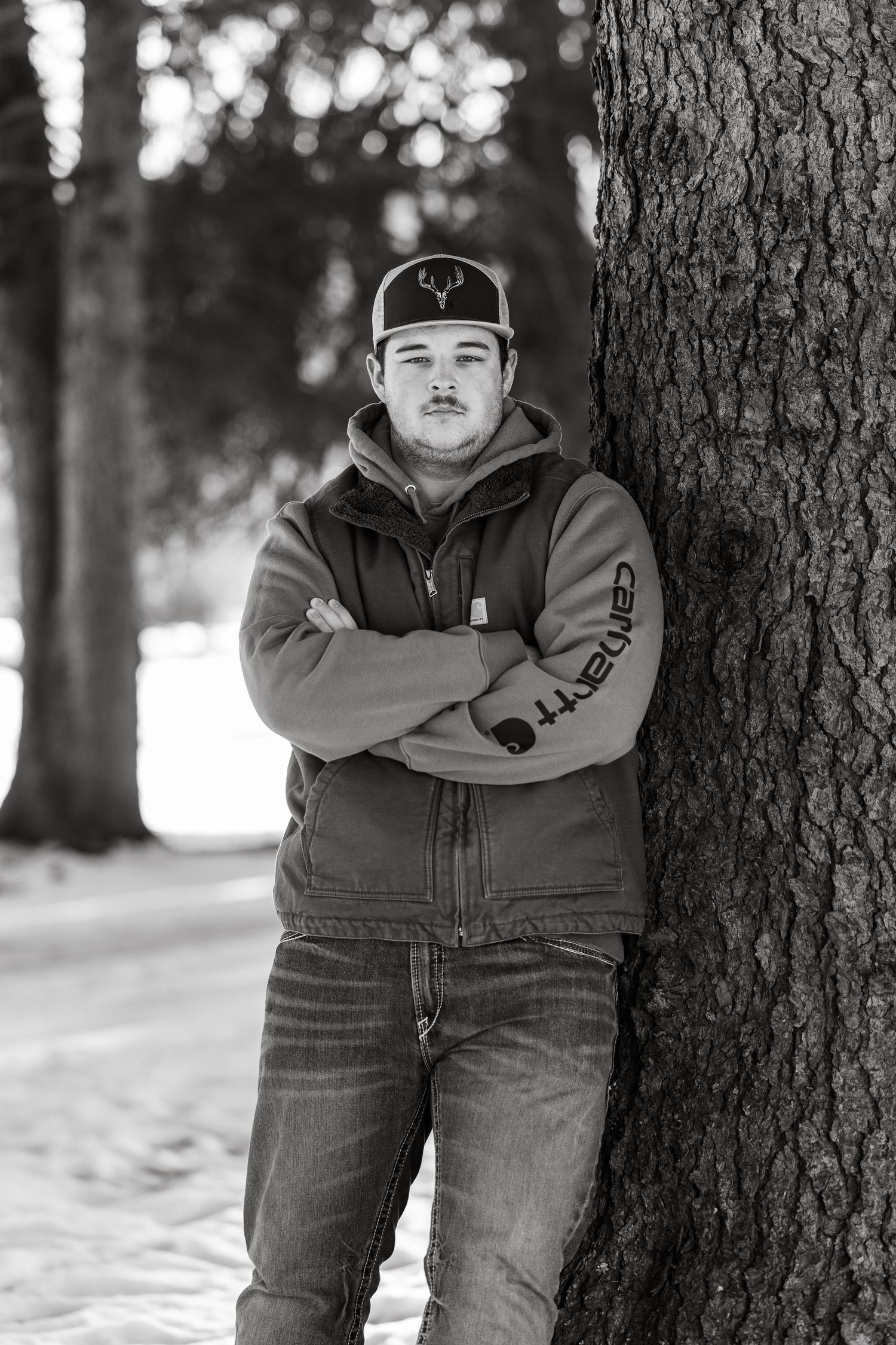 Person in work jacket and baseball cap leaning against tree trunk in winter setting.