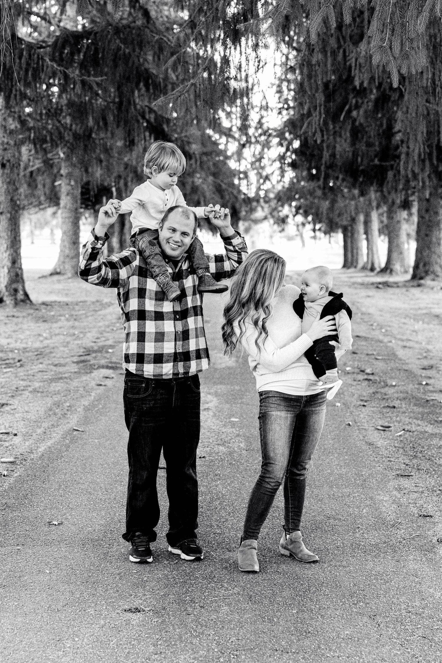 Family enjoying playful moment together on tree-lined path in black and white photo.