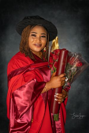 Graduate in red academic regalia holds a diploma and flowers during celebratory portrait session with moody lighting.