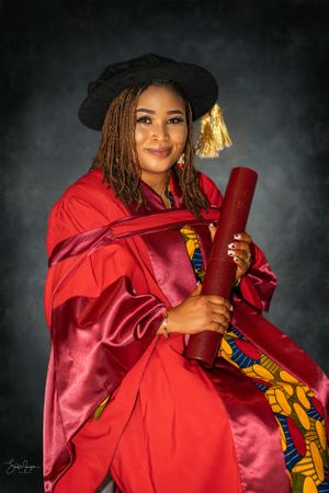 A graduate in red academic regalia and mortarboard with gold tassel holds their diploma against a dark background.