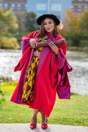 Graduate in bright red academic regalia and black cap poses outdoors with diploma on sunny autumn day.
