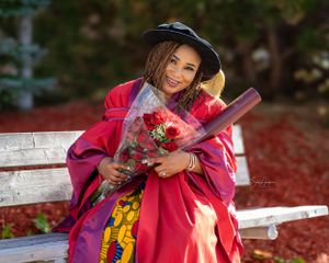 Graduate in red and pink academic regalia holds diploma and flowers while sitting on white bench in garden setting.
