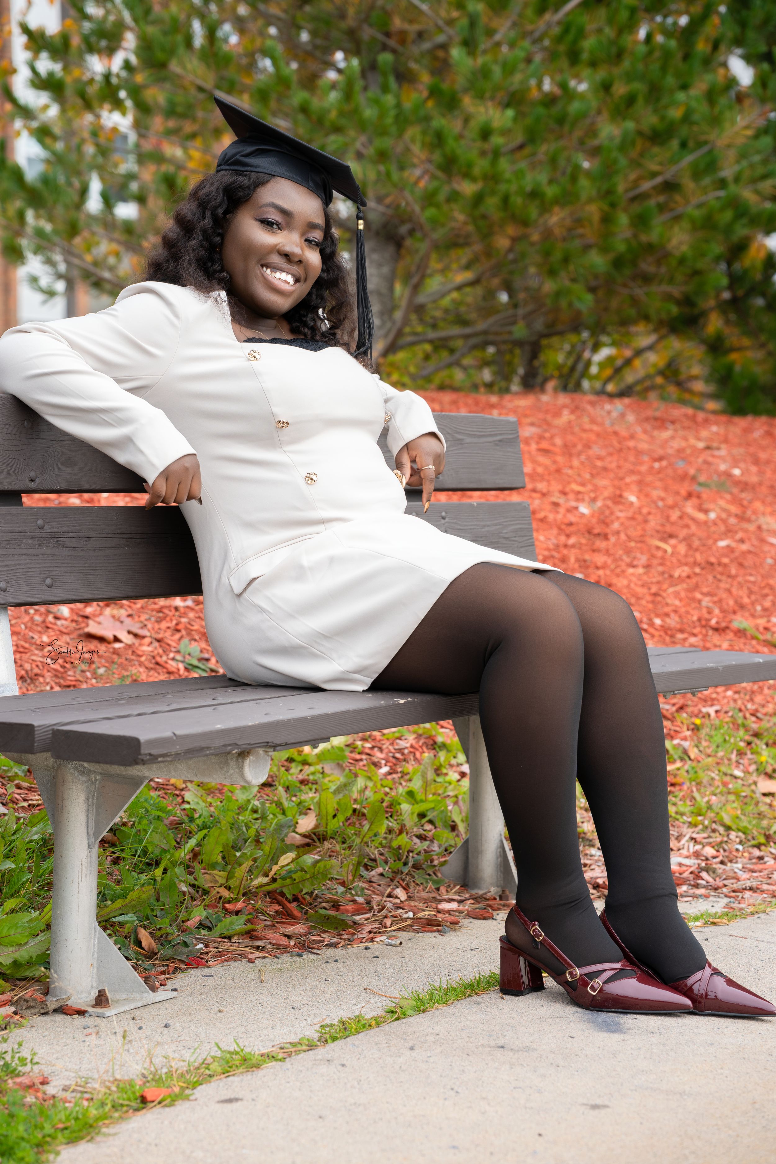 A person in a white dress and black tights lounges on a park bench surrounded by autumn leaves.