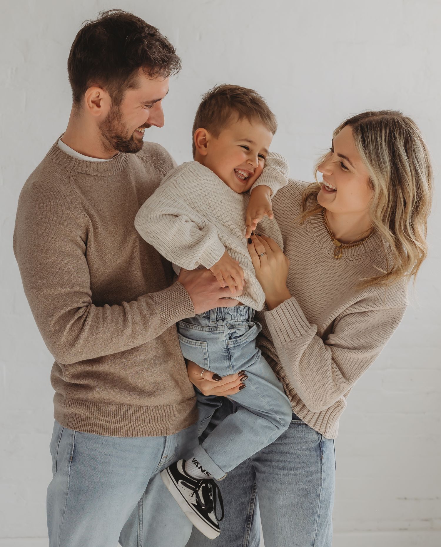Family photographer in Leicestershire. Happy family in matching beige sweaters share a joyful moment together against white background.