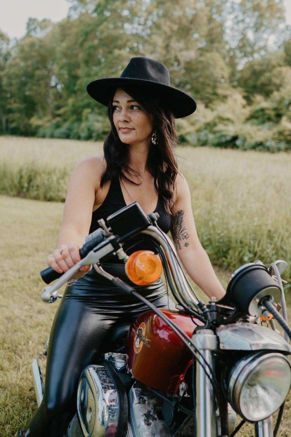 Person in black hat sits on vintage motorcycle in sunny meadow with golden autumn light filtering through trees.