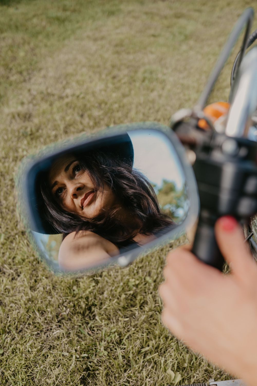 A motorcycle side mirror reflection shows dark hair blowing in the breeze against a grassy outdoor background.