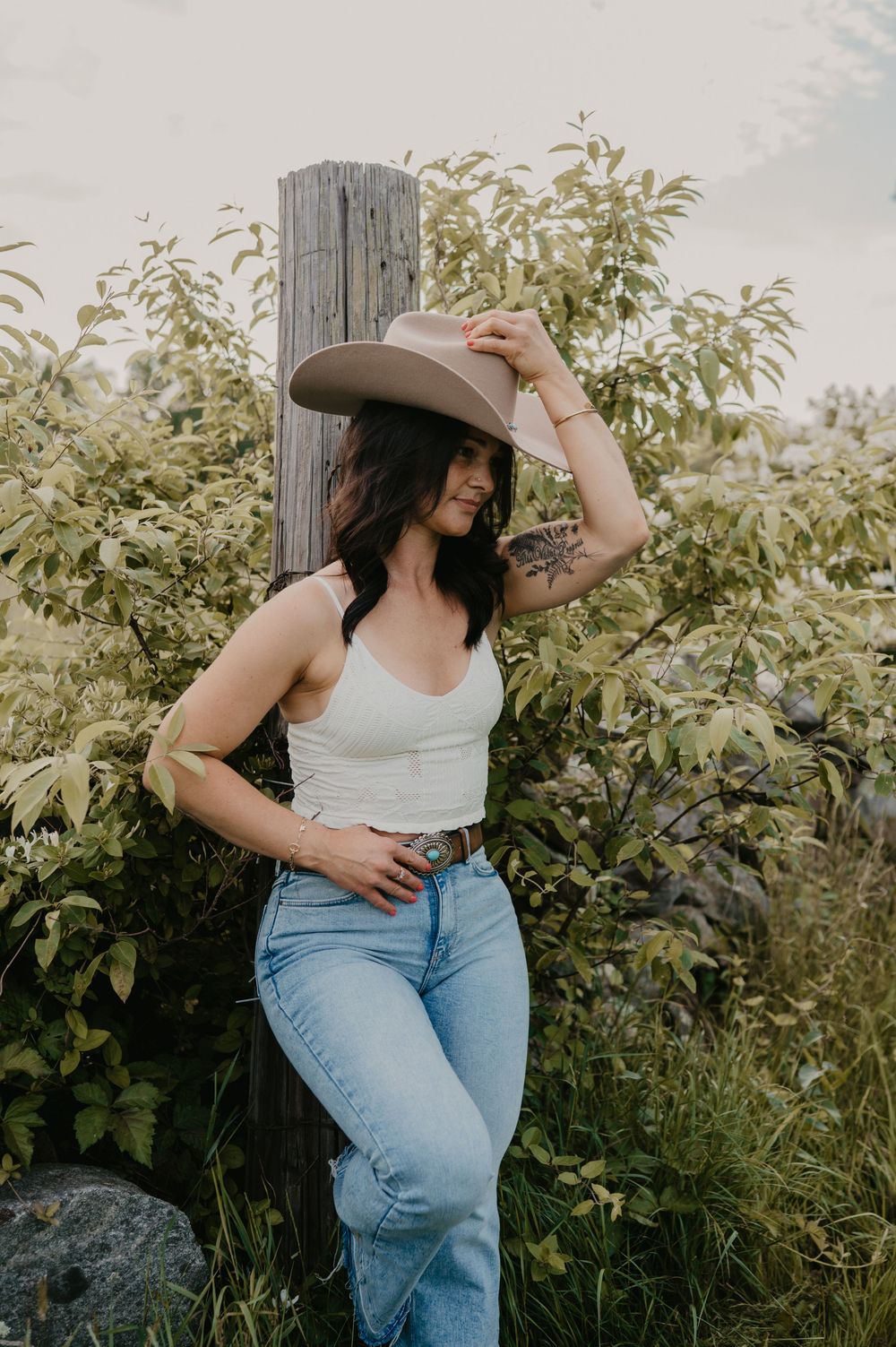A person in a white top and blue jeans poses gracefully against a natural backdrop with lush greenery.