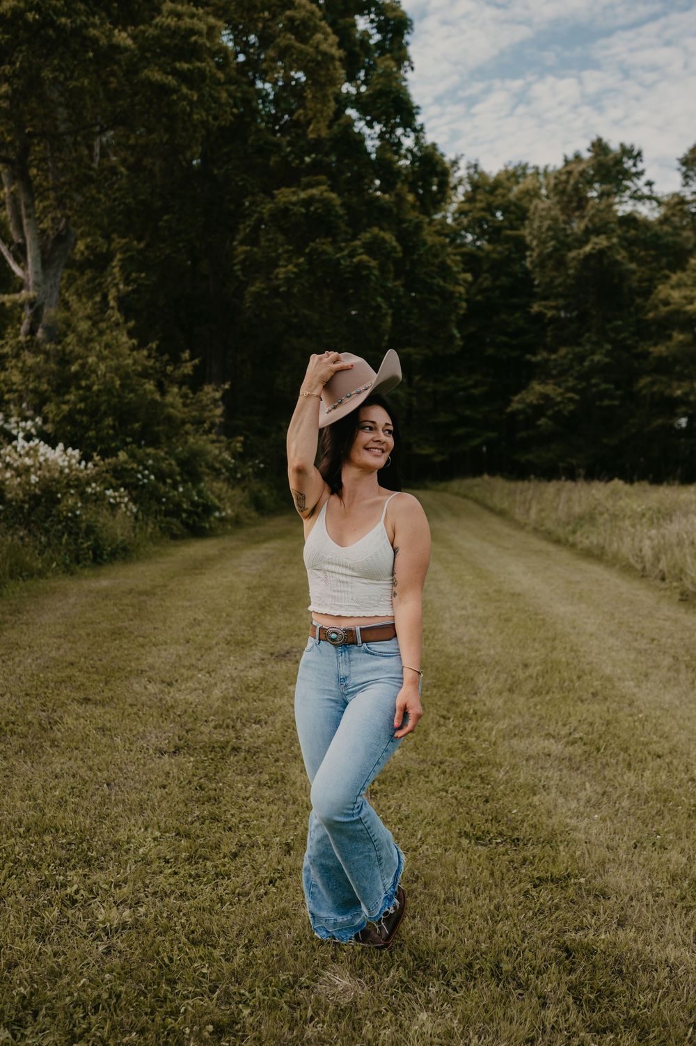 A figure in a white tank top, cowboy hat and jeans stands on a grassy path with trees in the background.