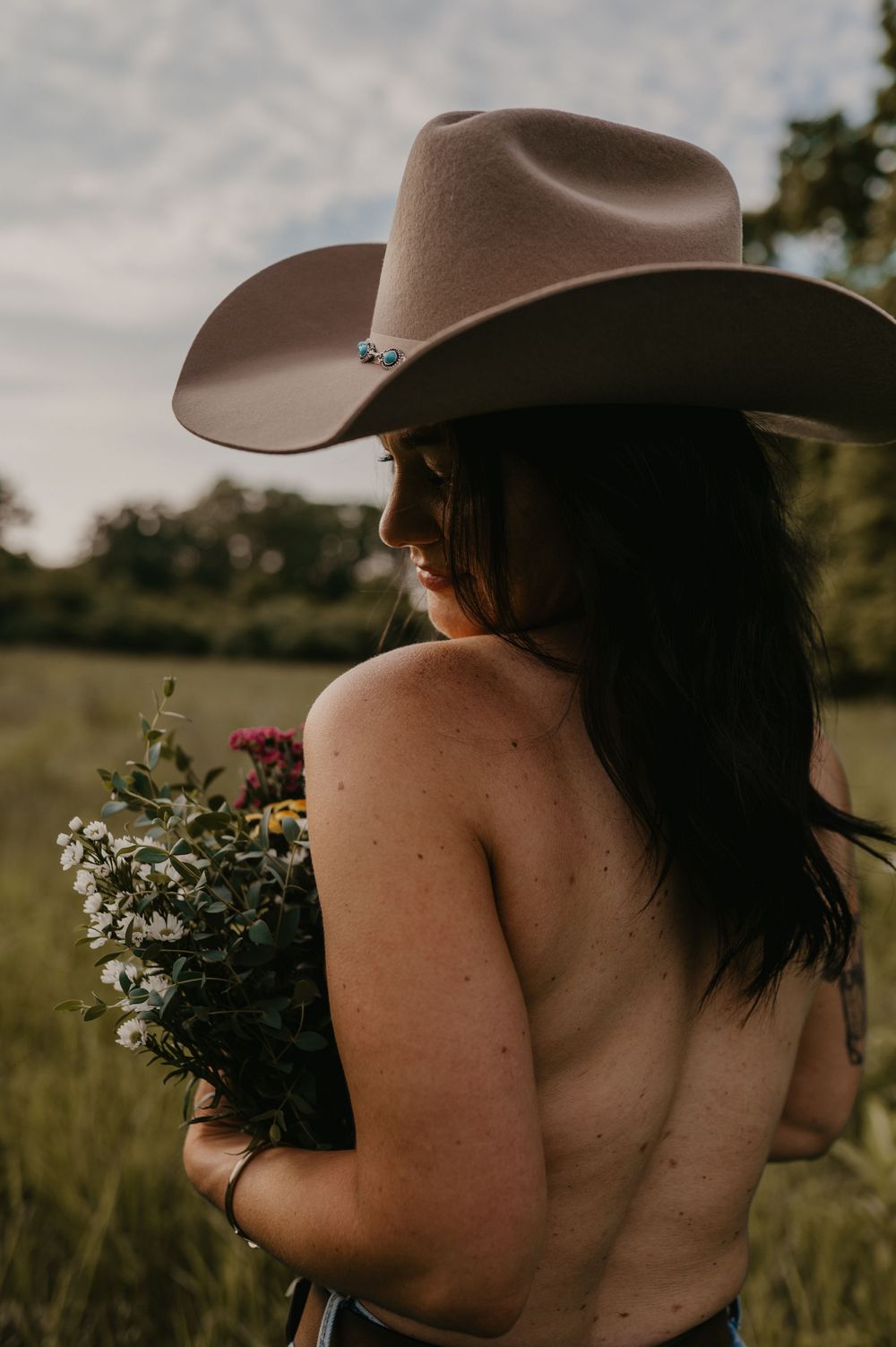 A person wearing a brown cowboy hat holds a bouquet of wildflowers while standing in a sunlit meadow at sunset.