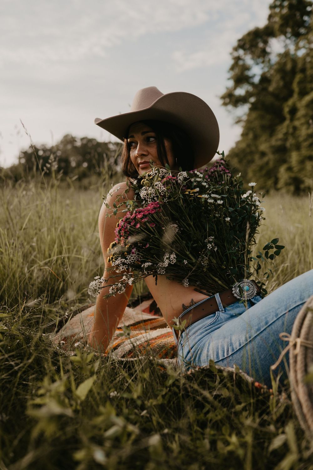 Someone in a cowboy hat sits in a grassy field holding a rustic bouquet of wildflowers and dried flowers.