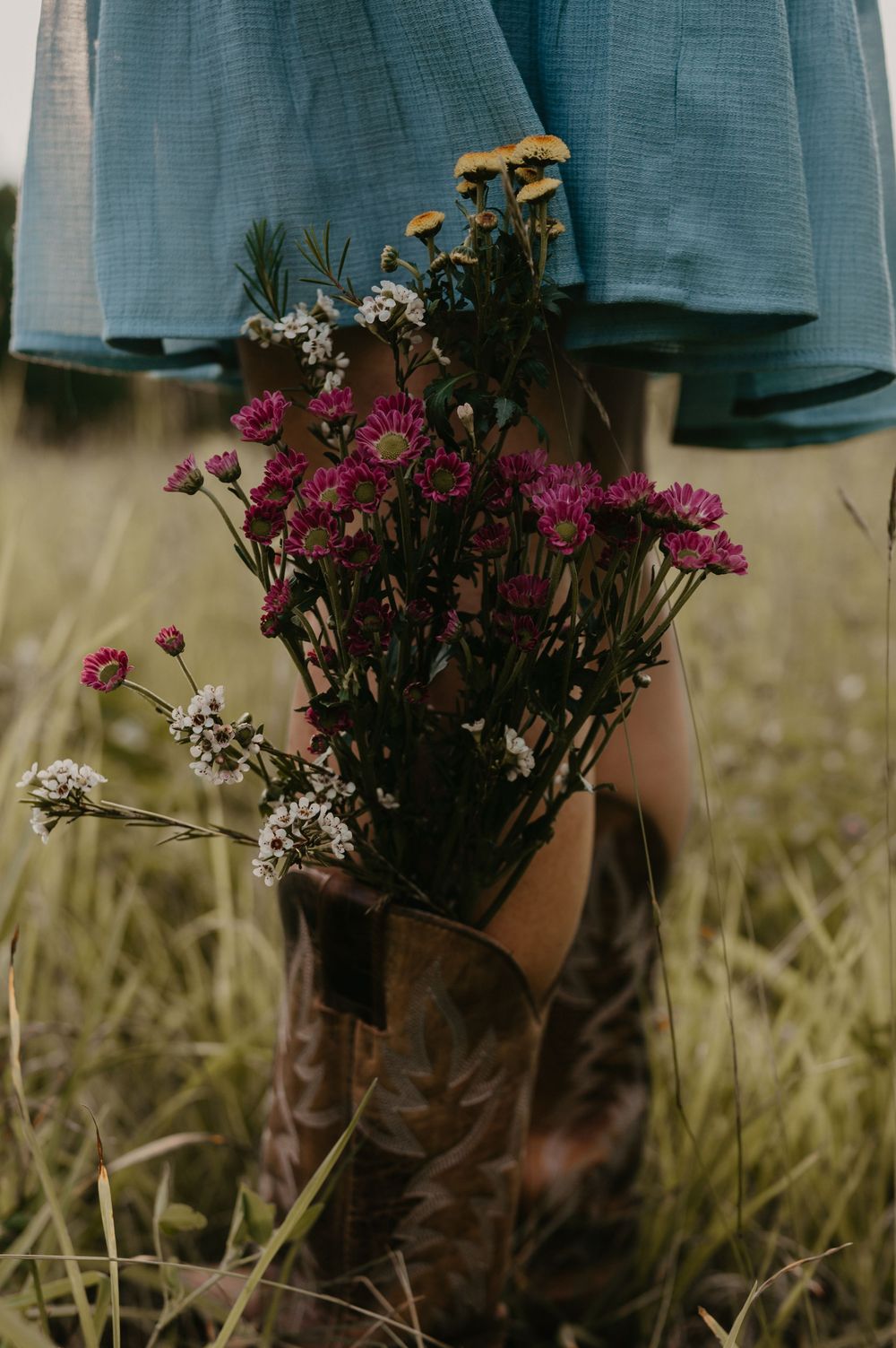 Pink and white wildflowers arranged in a brown leather cowboy boot against tall grass and blue denim.