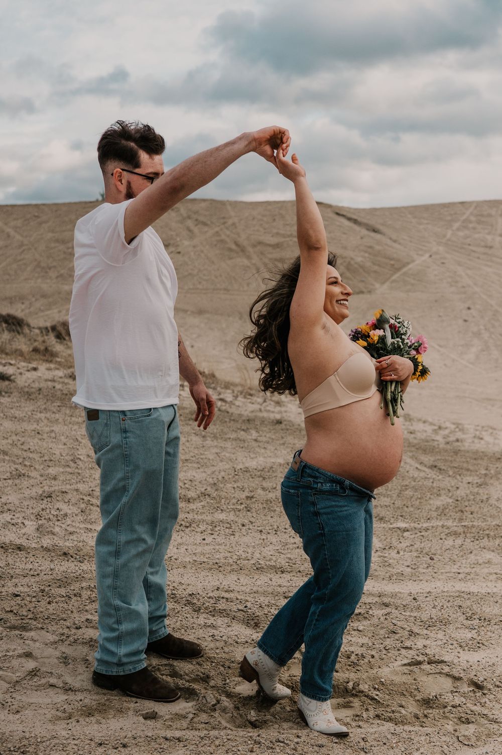 Expecting couple dances together in desert landscape with woman holding flowers against cloudy sky.