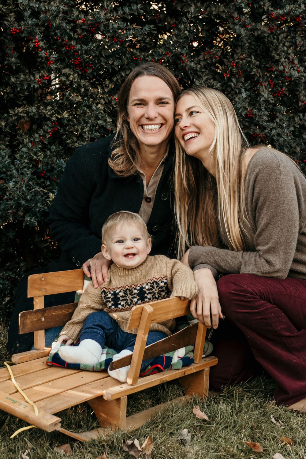 Family of three laughing together while sitting on wooden sled with red berries and greenery in background.