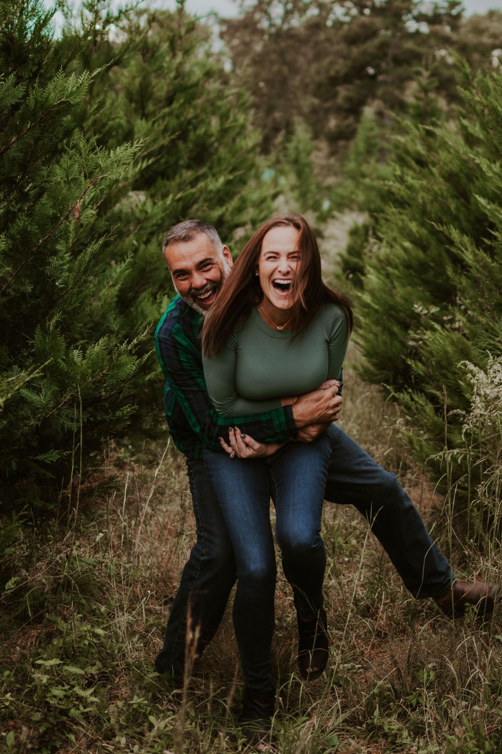 Laughing couple embraces outdoors surrounded by evergreen trees in a candid photoshoot series.