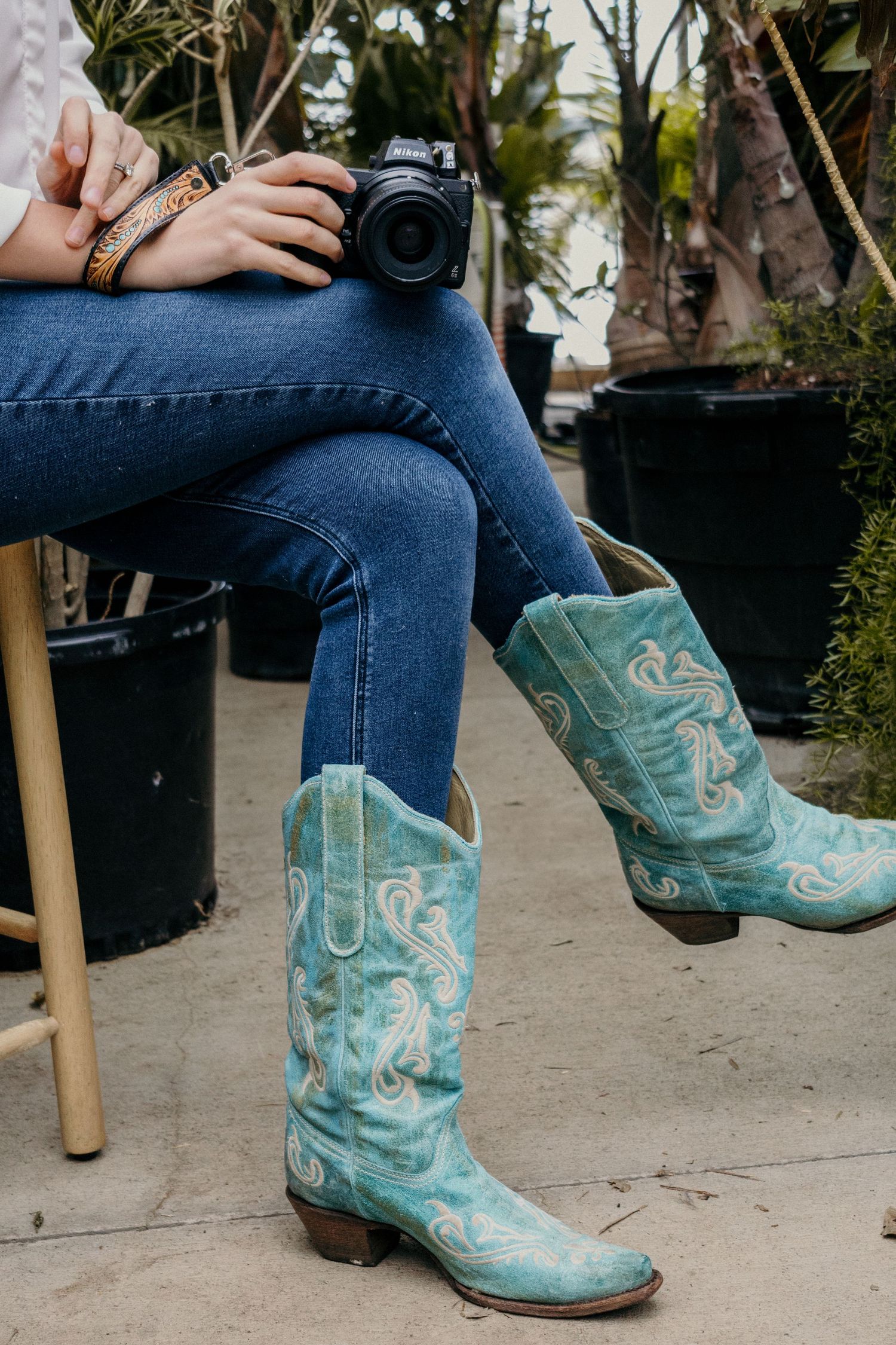 Someone wearing turquoise cowboy boots and blue jeans holds a camera while sitting on a wooden stool outdoors.