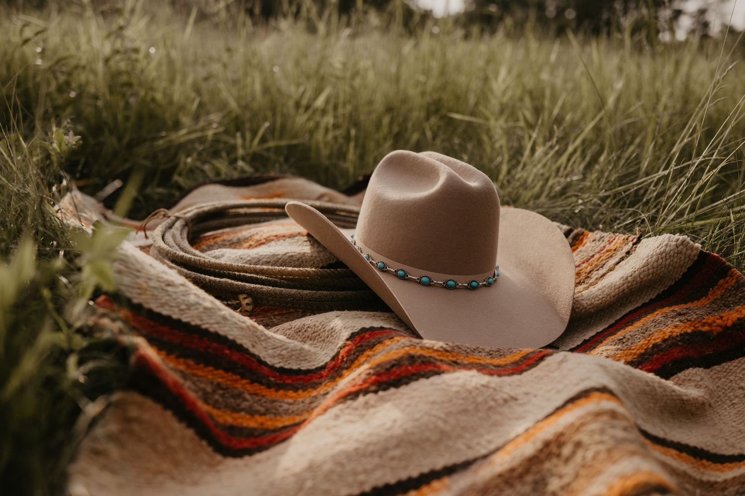 Brown cowboy hat with turquoise beaded band rests on southwestern style blanket in tall grass field.