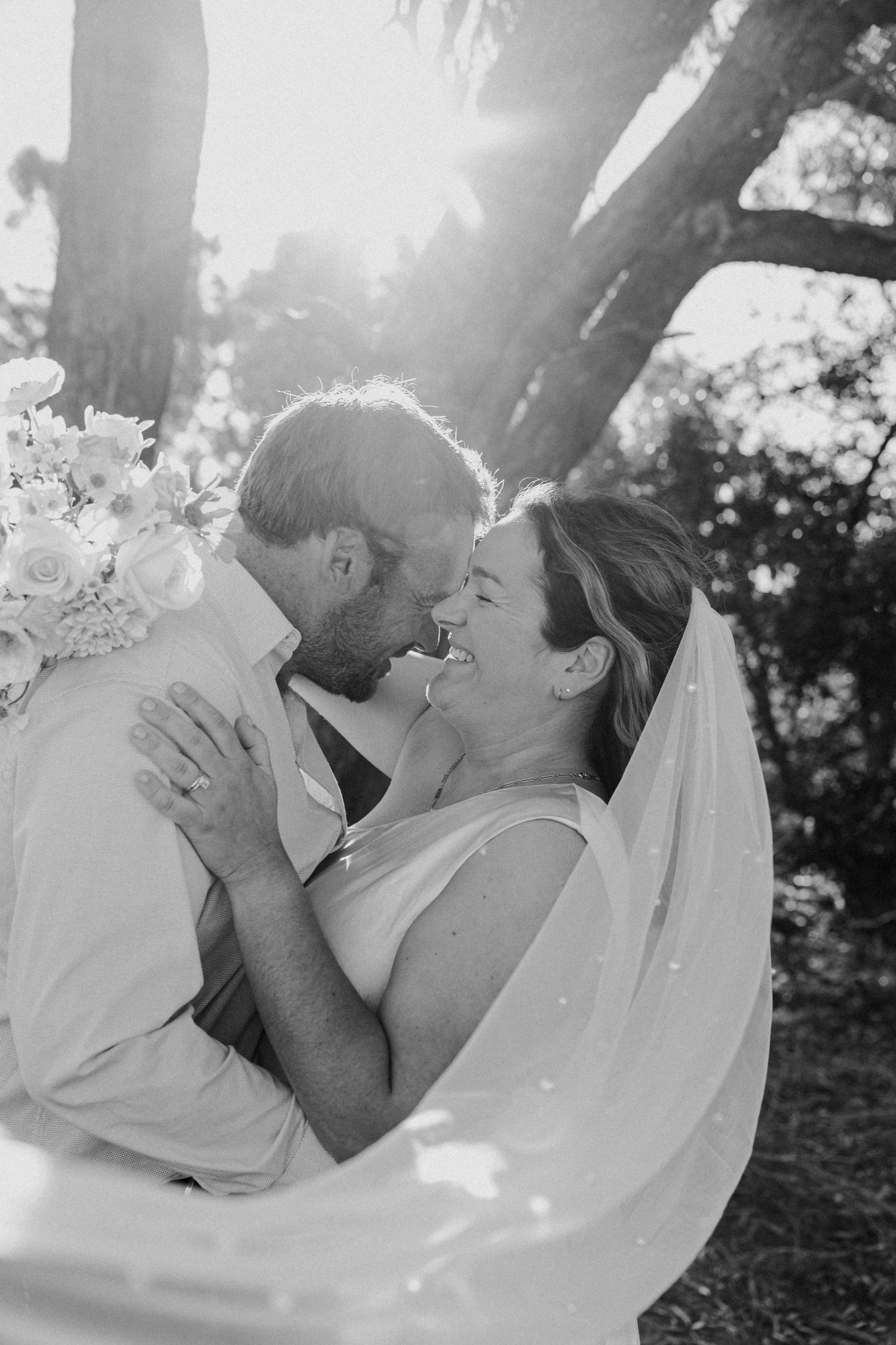 Romantic black and white wedding portrait with flowing veil and sunlight through trees.