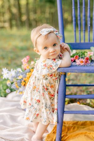 A baby girl in a floral dress and bow headband stands by a blue chair outdoors surrounded by colorful flowers.