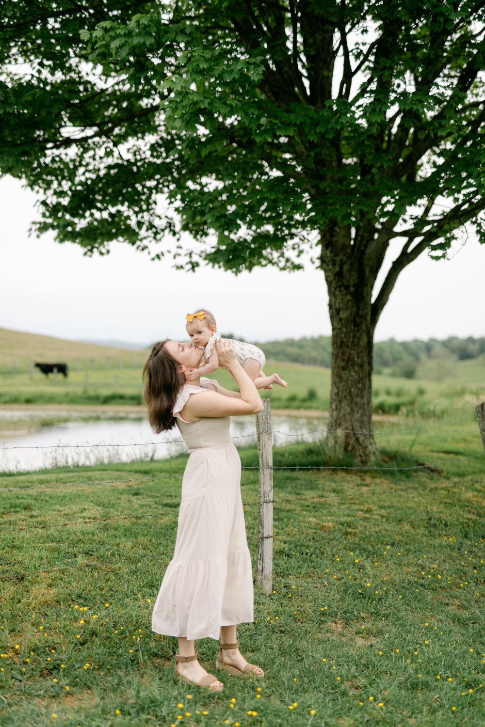 A mother in a white dress lifts her smiling baby up under a large tree by a pond.