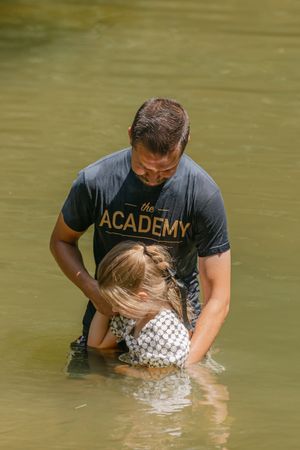 Man in Academy shirt baptizing a young child in a river outdoors.