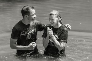 Two people standing in water during a baptism ceremony, both wearing black Academy shirts, in black and white.