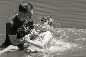 Man in Academy shirt baptizing a person in open water, captured in black and white with dramatic water splashes.