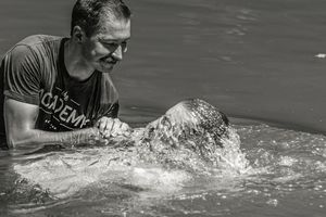 Man in t-shirt performing a water baptism ceremony, submerging a person in a river, black and white photo.