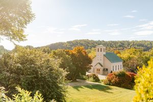 A white country chapel with green roof nestled among colorful autumn trees on a sunny hillside.