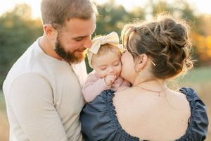 A couple shares a tender moment with their baby girl wearing a bow outdoors in warm golden light.