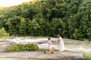 Romantic marriage proposal on rocky overlook above waterfall, lush green forest backdrop, man kneeling before woman in white dress.