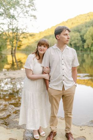 A couple in white outfits poses by a serene lake with lush green hills in the background during golden hour.
