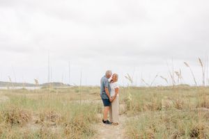 Couple embracing on a sandy beach path surrounded by tall sea grass under an overcast sky.