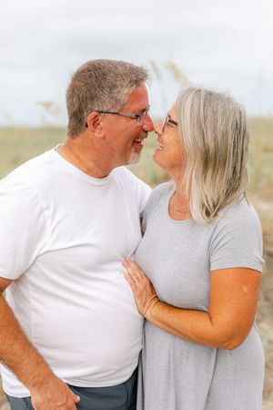 Older couple sharing a romantic moment outdoors, smiling and nearly kissing near sandy dunes.
