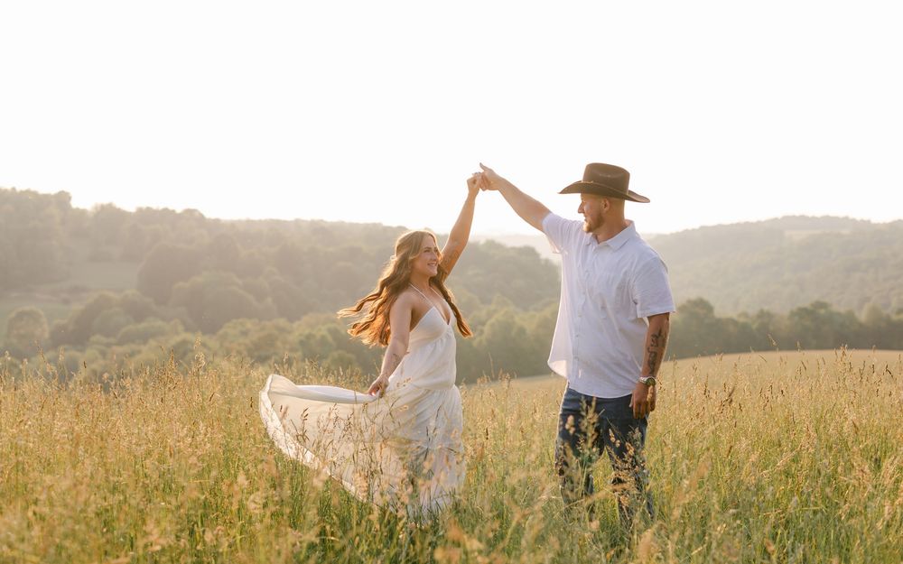 Couple dances in golden sunlit meadow, woman in white flowing dress spins joyfully.