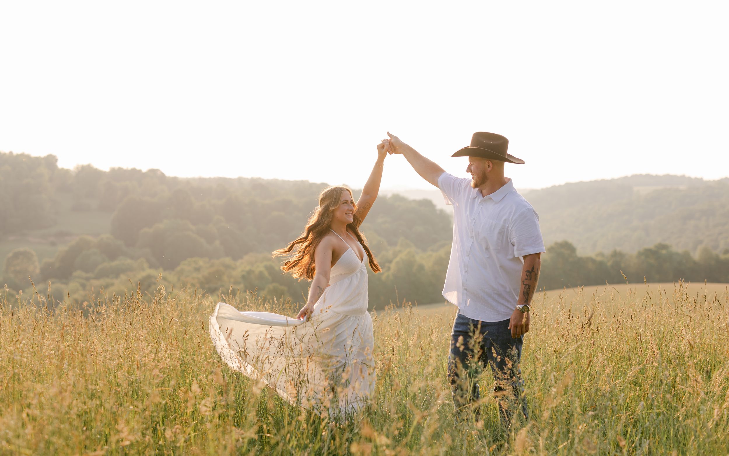 Couple dances in golden sunlit meadow, woman in white flowing dress spins joyfully.