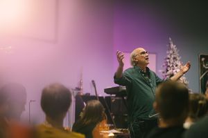 Elderly man in green shirt gesturing passionately while speaking to an audience in a dimly lit venue.